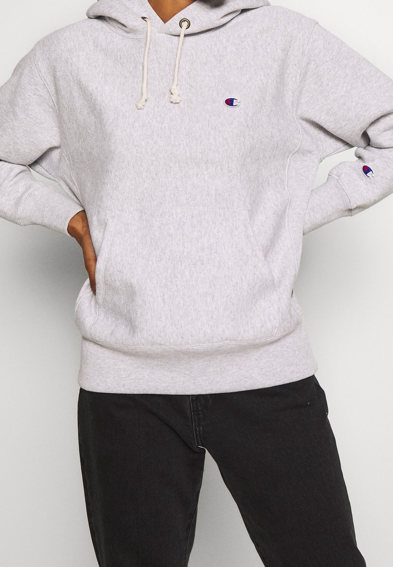 Personne portant un sweat à capuche Champion gris clair avec logo sur la poitrine et la manche, associé à un pantalon noir, mains posées sur les hanches.