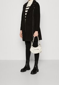 Svart ullblandad kappa med asymmetrisk dragkedjestängning, kombinerad med en randig polotröja, svarta leggings, kraftiga svarta stövlar och en liten vit handväska.