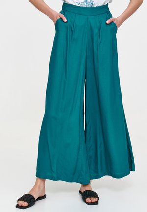 Turkusowe spodnie o szerokich nogawkach z zakładkami, wysokim paskiem w talii i bocznymi kieszeniami, wykonane z gładkiego materiału. Noszone z czarnymi plecionymi klapkami.