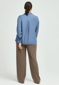 Blue long-sleeve blouse with a relaxed fit, back pleat detail, and button cuffs paired with brown and beige checked wide-leg pants.