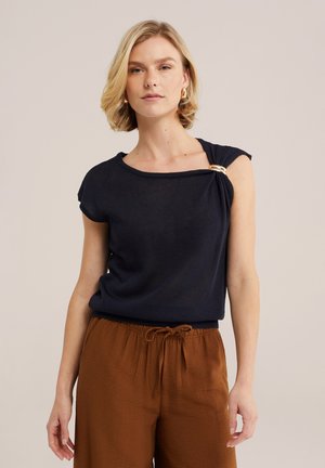 Vrouw met kort blond haar die een zwart mouwloos topje draagt met een gouden ringdetail op de schouder en bruine broek met trekkoord.