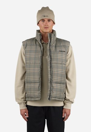 Padded vest with a checkered pattern in beige and dark green; features a high collar and front zipper. Worn over a ribbed long-sleeve shirt.