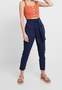 Top a tank cropped di un arancione ruggine con dettagli a bottoni, abbinato a pantaloni navy a vita alta con cintura annodabile. Sandali color cuoio completano l'outfit.