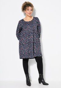 El vestido presenta un fondo azul marino con puntos florales rojos y blancos, mangas largas, un escote redondo, cintura fruncida y un corte holgado.