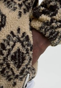 Beżowy polarowy materiał z czarnym geometrycznym wzorem, o teksturowanej, pluszowej powierzchni. Metka widoczna jest w pobliżu dolnego brzegu.