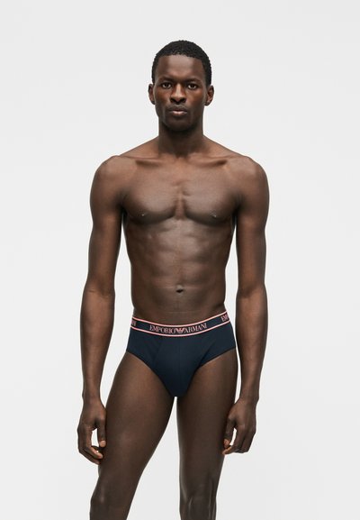 Άνδρας χωρίς πουκάμισο, όρθιος, φοράει μαύρα εσώρουχα Emporio Armani με ροζ γράμματα στη μέση, σε απλό ανοιχτό γκρι φόντο.