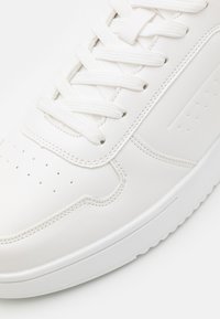 Weiße Ledersneaker mit texturiertem Obermaterial, flachen Schnürsenkeln, perforiertem Zehenbereich und Gummisohle, die ein sauberes Design und dezente Nähte aufweist.