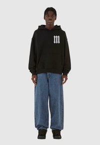Felpa nera con cappuccio e tasca frontale, logo bianco "m", abbinata a jeans a gamba larga blu e scarpe nere, in piedi su uno sfondo grigio.