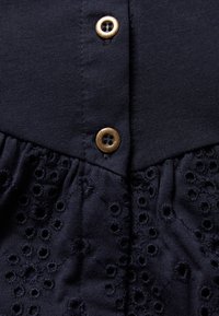 Námořnická modrá blůza s hladkou horní částí, detaily knoflíků a texturovanou dolní částí s vyšívanými očkovými vzory.