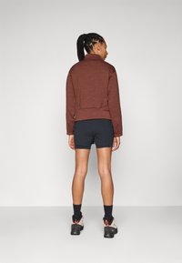 Brown, textured jacket with a high collar, paired with navy shorts. The model wears black socks and beige hiking boots with orange accents.