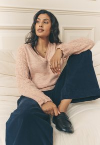 Jasnoróżowy dziergany sweter z dekoltem w serek i koronkowymi zdobieniami na rękawach, zestawiony z ciemnogranatowymi szerokimi spodniami sztruksowymi oraz czarnymi mokasynami.