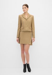 Tweed-kaksiosainen puku vaaleanruskeana, jossa on istuva bleiseri kultanapilla ja minihame sivuaukkoineen. Mustat nilkkurit täydentävät asun.