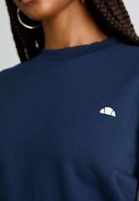 Sweatshirt bleu marine avec un col rond. Présente un petit logo blanc en forme de demi-lune sur la zone de la poitrine gauche. Texture lisse.