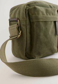Sac en toile vert olive avec fermeture éclair, présentant une bandoulière texturée et du matériel en laiton. Forme rectangulaire avec un détail de poche latérale.