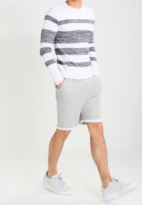 Homme portant un pull à rayures blanches et grises, un short gris clair et des baskets blanches, debout les mains dans les poches devant un fond uni.