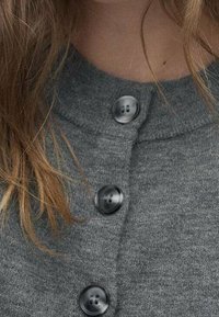 Grijze gebreide cardigan met een ronde halslijn en zwarte plastic knopen. Close-upweergave toont de zachte textuur en de knoopdetails.