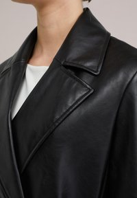 Svart läderjacka med bred krage, slät yta och skräddarsydd passform. Sedd från axelområdet, vilket framhäver designens detaljer.