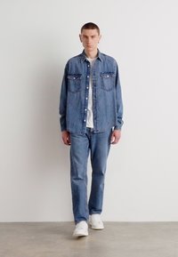 Spijkerhemd met twee borstzakken, gecombineerd met bijpassende spijkerbroek. Witte t-shirt eronder, afgemaakt met witte sneakers.