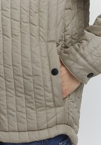 Giacca trapuntata beige con una tasca laterale con bottone a scatto nero. Il tessuto ha una texture liscia e motivi di cucitura orizzontali.