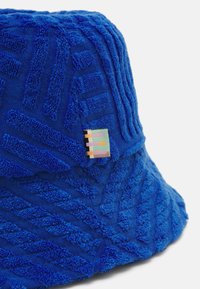 Chapeau seau en velours bleu avec un motif rayé texturé et une étiquette colorée. Tissu doux avec un sommet arrondi et un large bord évasé.