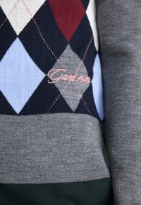 Grå genser med et argyle-mønster i marineblå, rød, lyseblå og grå. Har rosa brodert tekst "Gant 1949" nær bunnkanten.