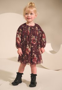 Ziedu kleita burgundijas krāsā ar garām, pūstām piedurknēm un sapinumu pie kakla; valkāta ar melnām lakotām mežģīņu zābakiem ar volānu akcentiem.