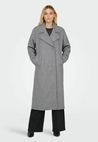 Lungo cappotto grigio realizzato in tessuto morbido, con collo a cono, tasche laterali e una silhouette dritta, abbinato a pantaloni neri a gamba larga.