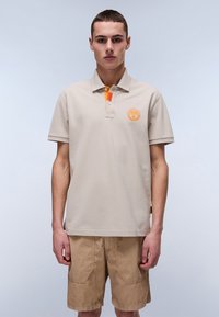 Beige polo t-shirt med en tekstureret overflade, orange kraveaccent og rundt logo på brystet. Bæres med beige shorts med lommer.