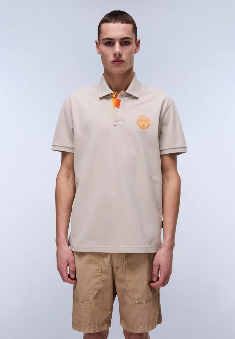 Beige polo t-shirt med en tekstureret overflade, orange kraveaccent og rundt logo på brystet. Bæres med beige shorts med lommer.
