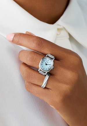 Mano con pelle marrone che indossa un orologio ad anello d'argento al dito medio e un anello d'argento intrecciato all'anulare su tessuto bianco.