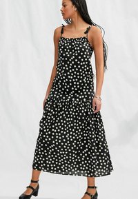 Abito maxi nero con pois, dotato di una gonna a strati, con scollatura arricciata e spalline da annodare. Realizzato in tessuto leggero con una texture liscia.