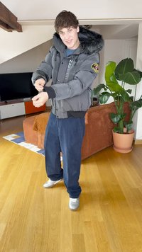 Μεταλλαχτη αναδιπλούμενη τζακετα σε σκούρο γκρι με κουκούλα επενδεδυμένη με γούνα, μαύρα μανίκια και λεπτομέρειες με λογότυπο. Φοριέται με ναυτικές βερμούδες από καφέ κορντουρόι και λευκά αθλητικά παπούτσια. Ξύλινο πάτωμα.