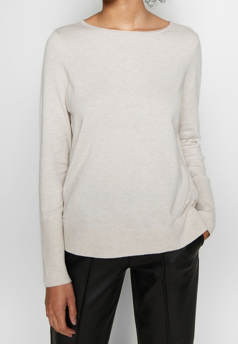 Femme portant un pull à manches longues beige clair et un pantalon en cuir noir, une main dans la poche, sur un fond uni.