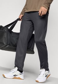 Pelēkie sporta bikses ar ciešu siluetu, kas izceltas ar gludu tekstūru un elastīgu jostasvietu. Saskaņotas ar baltām čībām un melnu sporta somu.