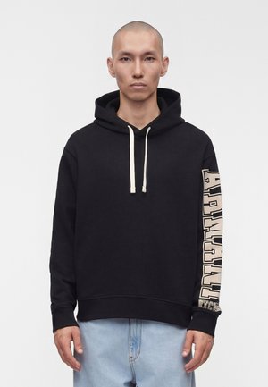 Mand med barberet hoved iført en sort hættetrøje med teksten "ARMADA" på ærmet og lyseblå jeans, stående mod en ensfarvet baggrund.