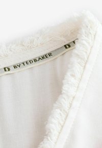 Valge riidekangas tekstuuriga frantsiisiga kaeluses. Silt on tume roheline ja seda on kirjas "BYTEDBAKER". Materjal näeb välja pehme ja kerge.