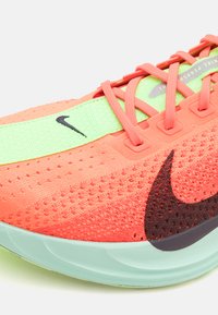 Chaussure de sport avec une tige en maille corail respirante, une bande de semelle intermédiaire vert néon et un logo swoosh noir. Surfaces texturées et contraste de couleurs vives.