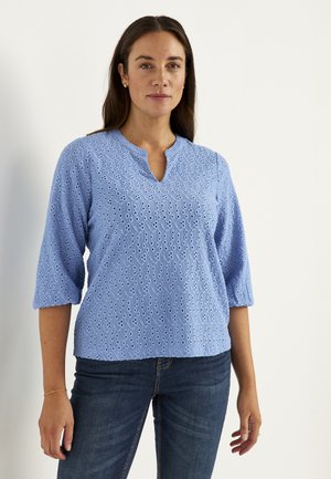 Kvinde med langt mørkt hår iført en lyseblå bluse med hulmønster og trekvartærmer og mørke jeans, står foran en ensfarvet baggrund.