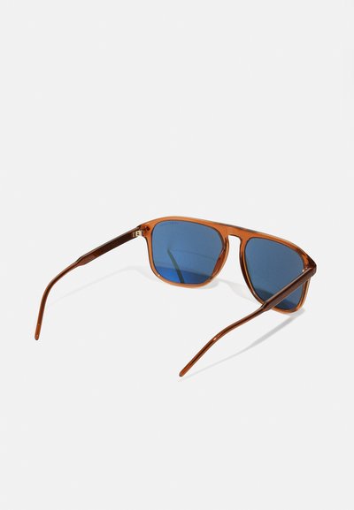 Lunettes de soleil en plastique marron avec des verres bleus, de forme carrée et avec des branches fines. Présentent des détails subtils sur la monture intérieure.