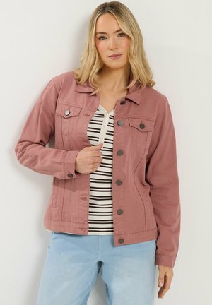 Mujer con cabello rubio que lleva una chaqueta rosa polvo abotonada sobre una camisa a rayas y vaqueros azul claro, posando contra una pared blanca.