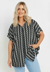 Camicia a maniche corte bianca e nera con motivo a onde, colletto, chiusura con bottoni e vestibilità rilassata, abbinata a jeans azzurri chiari.