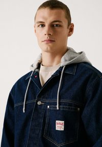Chaqueta de mezclilla oscura con capucha gris, botones plateados, bolsillo en el pecho con una etiqueta de Red Bull y detalles de costura prominentes.