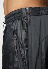 Shorts de sport noirs en tissu léger et texturé. Comprend une taille élastique, des poches zippées et des coutures rouges en accent.