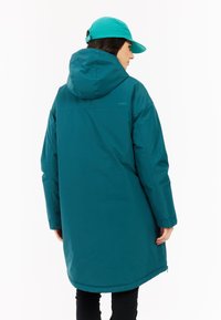 Abrigo largo impermeable de color azul verdoso con capucha, con cierre de cremallera y bolsillos laterales, llevado con una gorra a juego color turquesa. Textura suave.