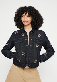 Rifľová bunda tmavomodrej farby s vyšitými erbmi leva v zlatej a červenej farbe. Obsahuje zapínanie na gombíky a detail na náprsnej vrecku.