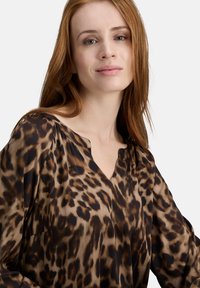 Bluse im Leopardenmuster mit weichem, seidigem Material, V-Ausschnitt und langen Ärmeln. Vorherrschende Farben sind Braun und Schwarz mit subtilen Kontrasten.