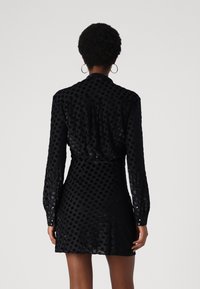 Robe noire avec un motif en points texturé, des manches longues et une taille cintrée. Le tissu a un léger brillant et une jupe évasée.