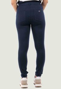 Pantaloni stretch blu navy con una texture liscia, vita alta e due tasche posteriori. Indossati con scarpe da ginnastica di colore chiaro.