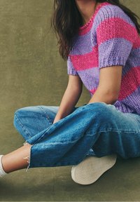 Persona sentada con las piernas cruzadas, vistiendo un suéter de punto a rayas púrpura y rosa, jeans azules con bordes deshilachados, calcetines blancos y zapatillas.