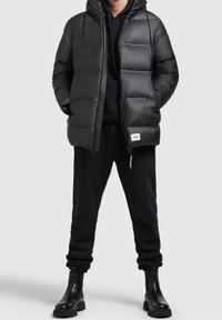 Schwarze Steppjacke mit Kapuze, gestepptem Muster, Fronttaschen und Reißverschluss, kombiniert mit schwarzen Hosen und Knöchelstiefeln.